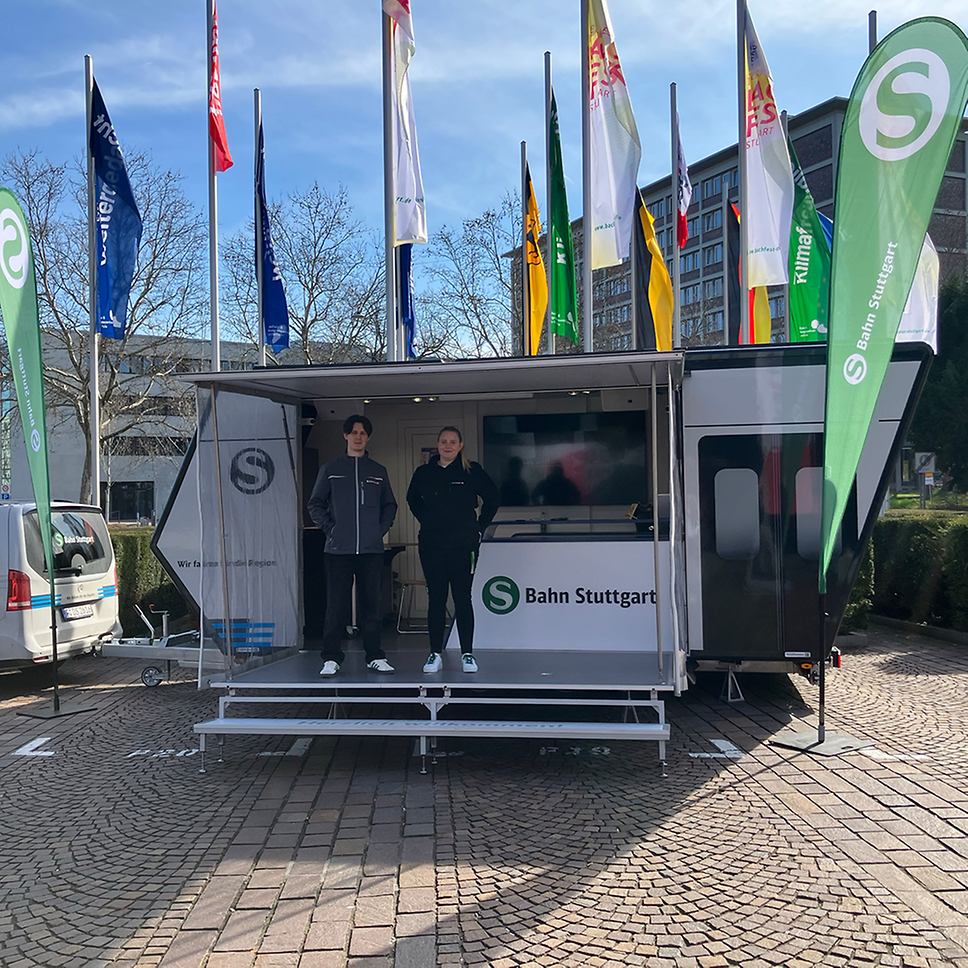 Zwei Kollegen der S-Bahn Stuttgart stehen vor dem Infomobil und lachen in die Kamera.