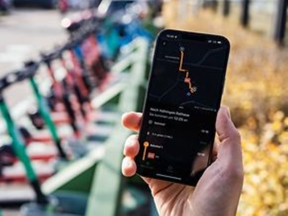 Ein Handy, auf dem die Mobility Stuttgart App angezeigt wird. Im Hintergrund sind E-Roller zu erkennen.